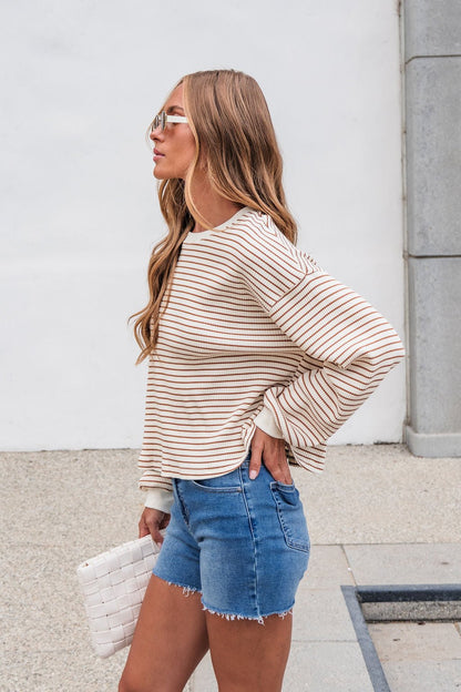 Woman in the Sutton Striped Pullover - Cream with denim shorts stands outside, holding a white clutch, facing left.