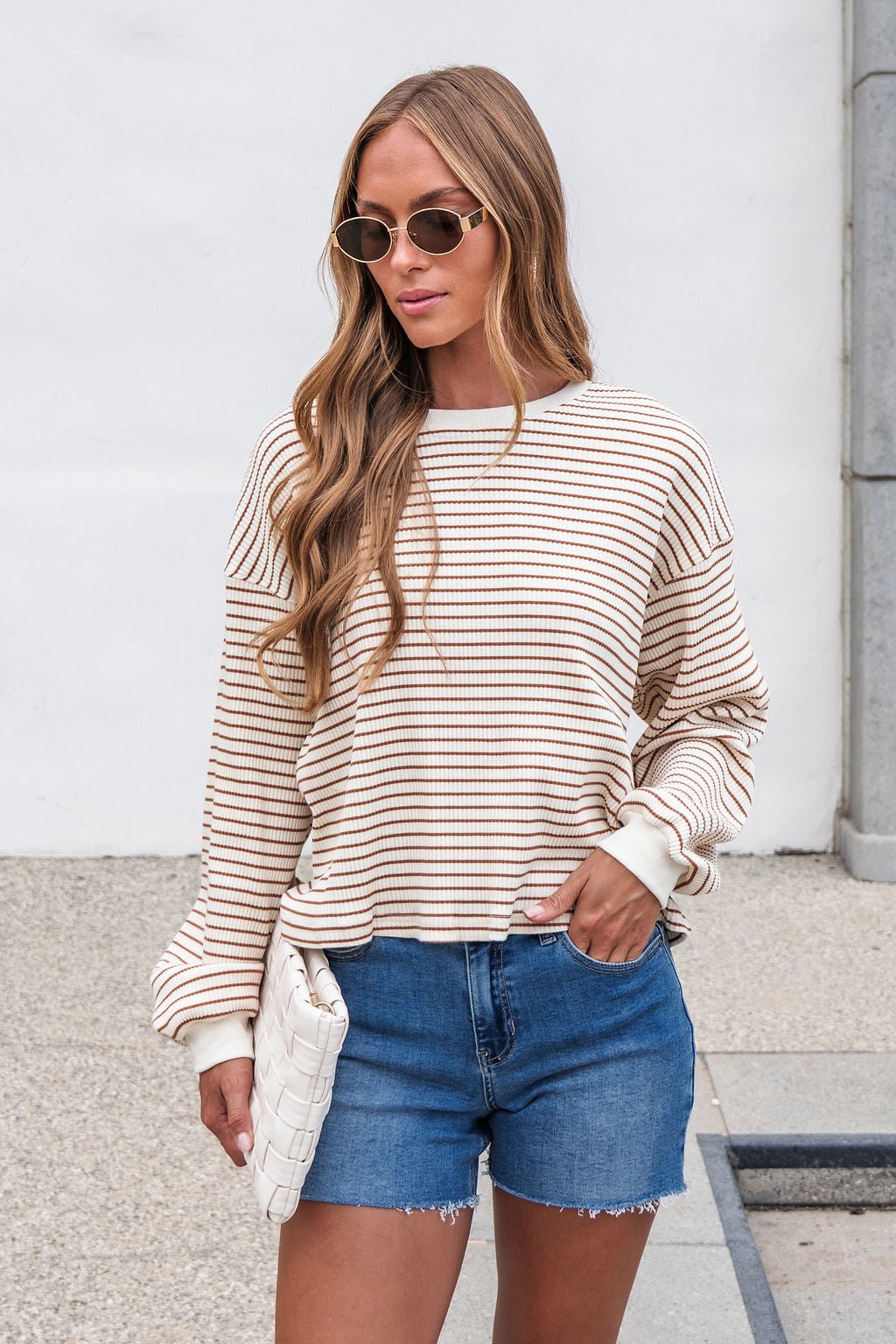 Woman in sunglasses wears the Sutton Striped Pullover - Cream and denim shorts, holding a white clutch outdoors.