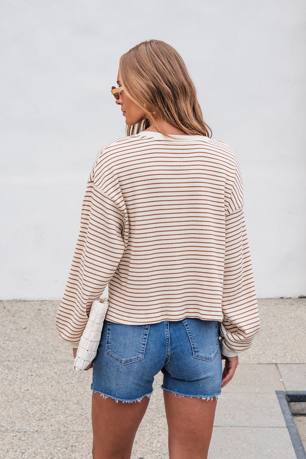 Woman with wavy hair, wearing the Sutton Striped Pullover - Cream and denim shorts, stands outdoors holding a white clutch.