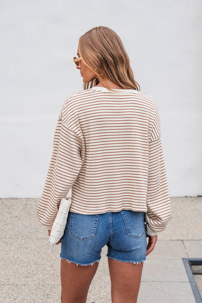 Woman with wavy hair, wearing the Sutton Striped Pullover - Cream and denim shorts, stands outdoors holding a white clutch.