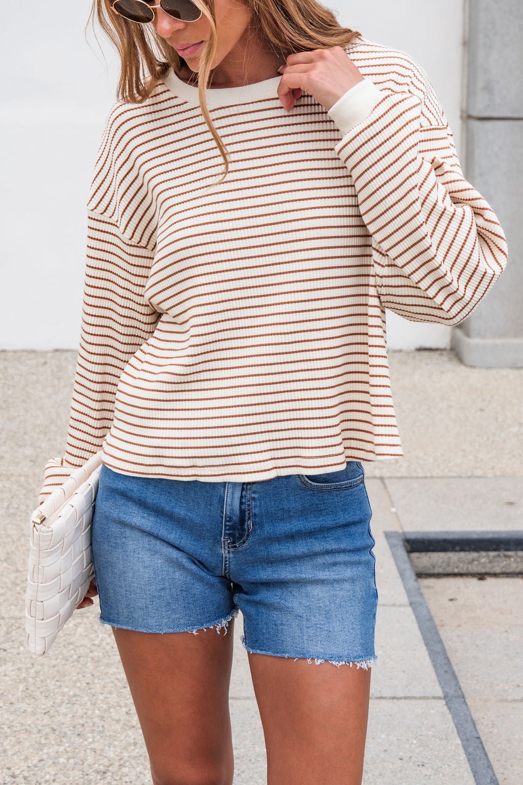Woman in sunglasses, Sutton Striped Pullover - Cream, blue denim shorts, holding a white woven clutch outdoors.