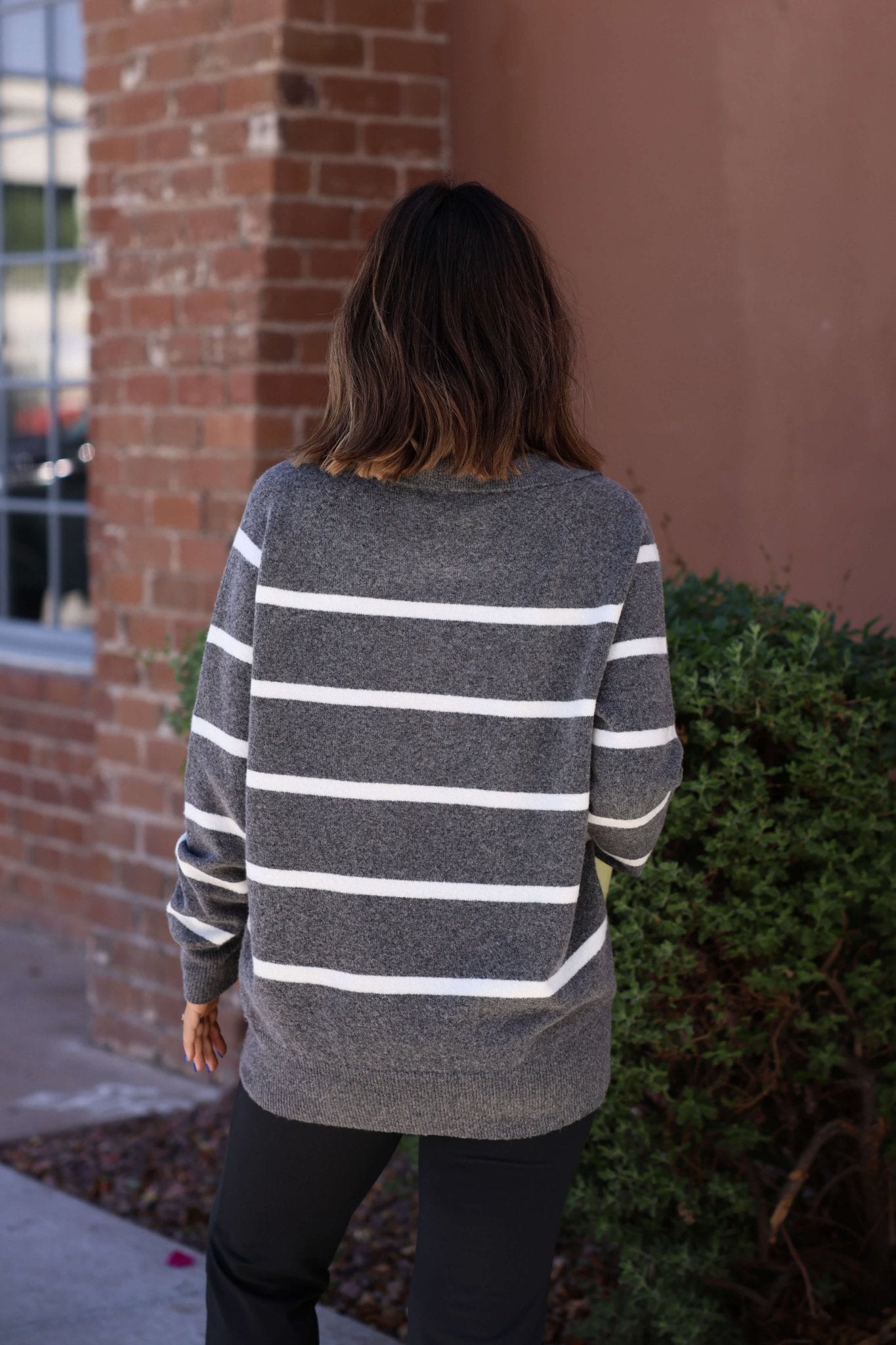 Wearing the Suzanne Grey & White Striped Sweater, a person with shoulder-length brown hair stands outside, back to the camera.