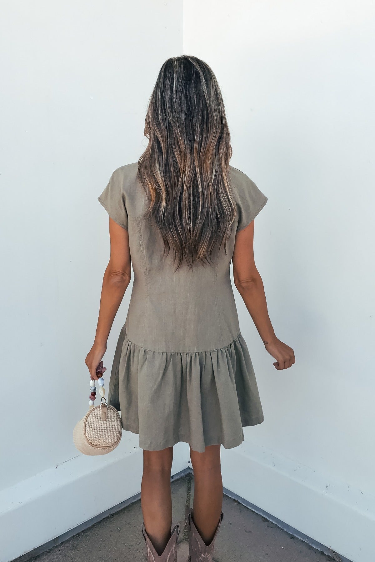 A woman in the Sweet Escape Linen Mini Dress - Olive and cowboy boots faces a white wall, holding a round woven handbag.