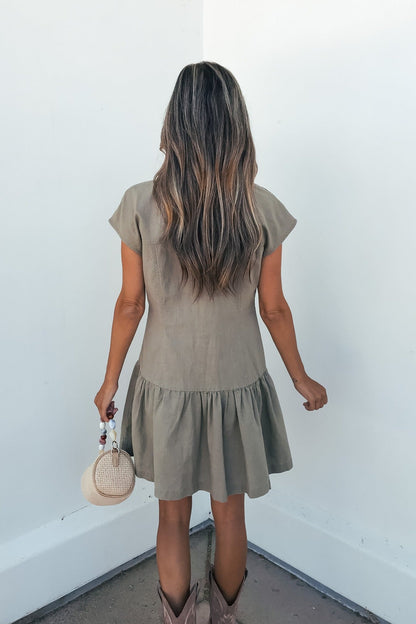 A woman in the Sweet Escape Linen Mini Dress - Olive and cowboy boots faces a white wall, holding a round woven handbag.