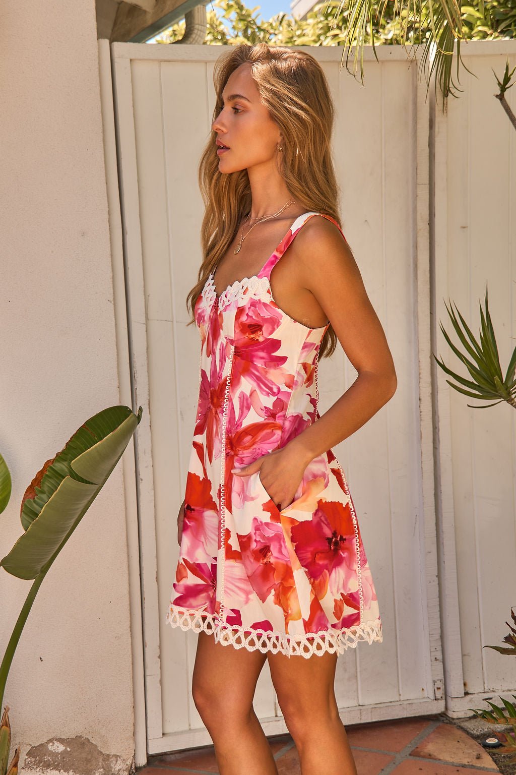 Woman in the Sweet Orchard Print Mini Dress - Pink stands outside near a white fence and tropical plants.