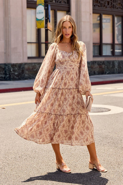 Woman in the Sweetheart Floral Print Midi Dress - Sand and sandals stands on a city street holding a clutch, buildings behind her.