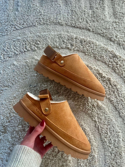 A hand holds a Tan Sherpa Lined Slipper - FINAL SALE, with fuzzy lining, on a textured beige rug.