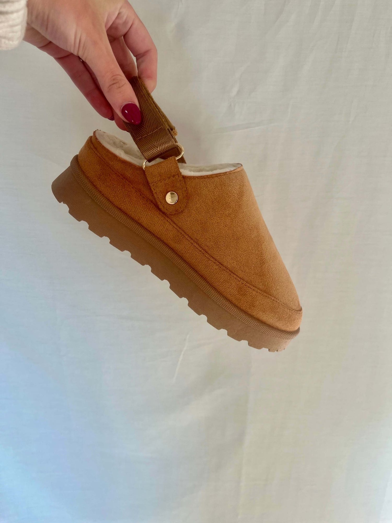 A hand with red nails holds a Tan Sherpa Lined Slipper with a back strap and textured sole against a plain light background.