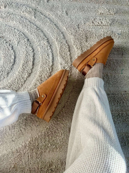 Person wearing Tan Sherpa Lined Slippers - FINAL SALE and light knit pants, seated on a beige and white circular rug.