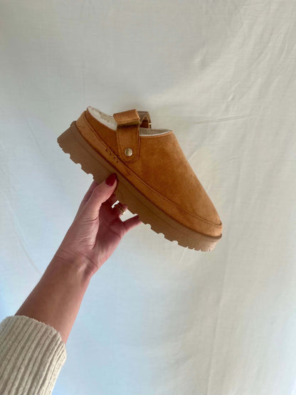 A hand holds up the Tan Sherpa Lined Slippers - FINAL SALE, featuring a thick sole and strap, against a light fabric background.