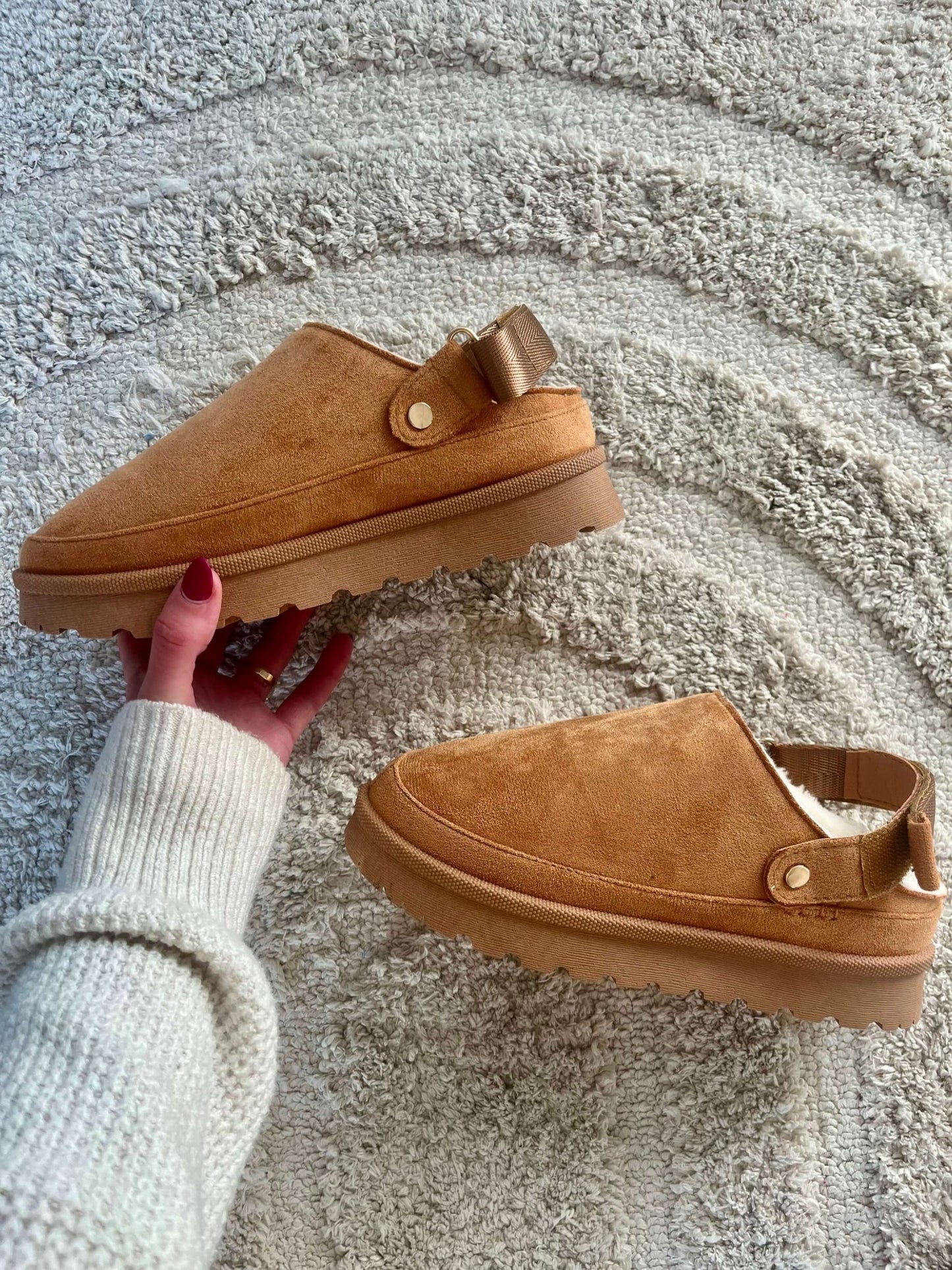 A hand holds Tan Sherpa Lined Slippers - FINAL SALE, showing their cozy slip-on style on a textured beige rug.