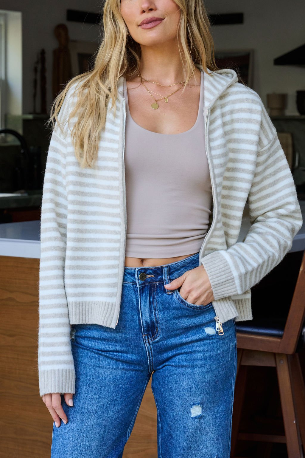 A woman in a Tan Striped Hooded Zip Up Jacket, beige tank top, and blue jeans stands in a modern kitchen with one hand in her pocket.