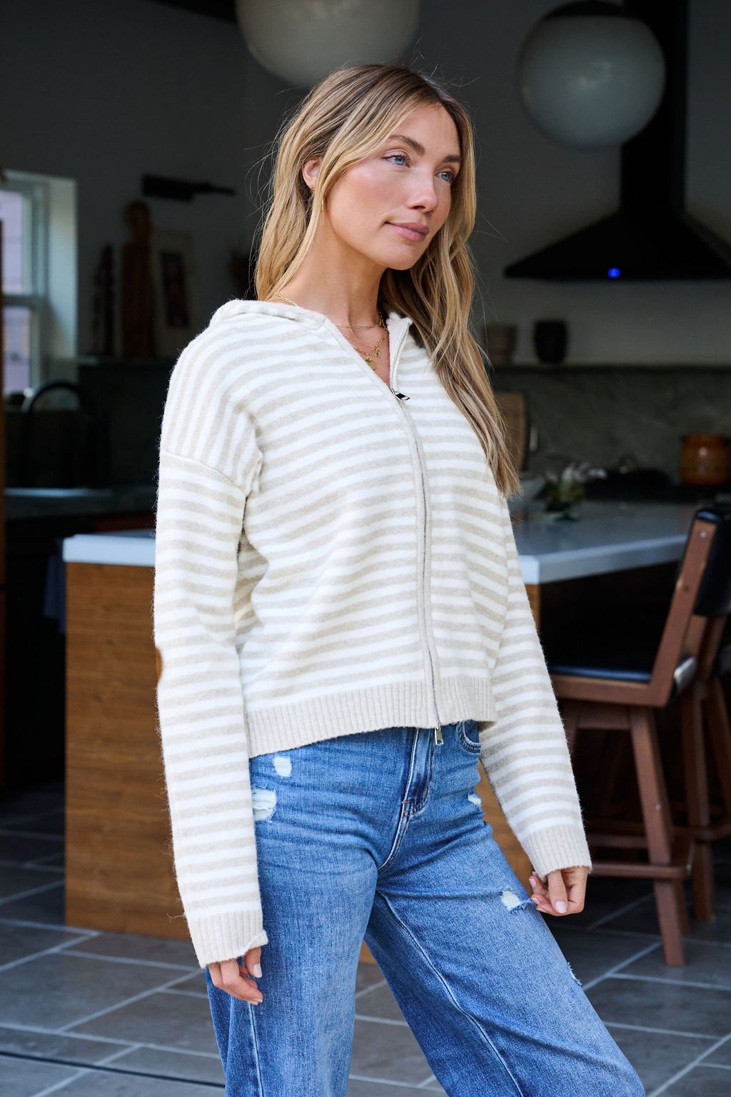 A woman wearing a Tan Striped Hooded Zip Up Jacket and blue jeans stands in a modern kitchen.