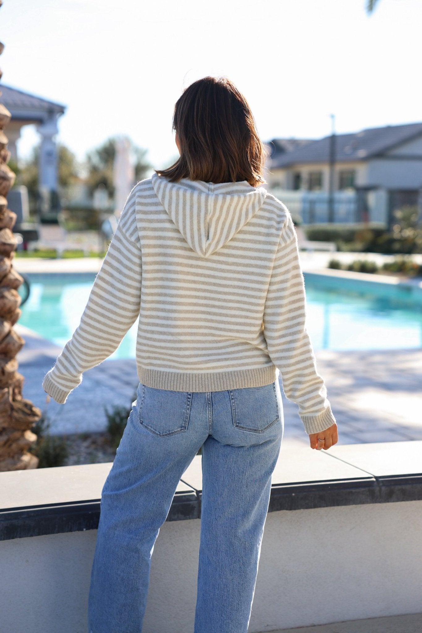 Someone wearing the Tan Striped Hooded Zip Up Jacket and jeans stands outdoors by a pool, facing away from the camera.