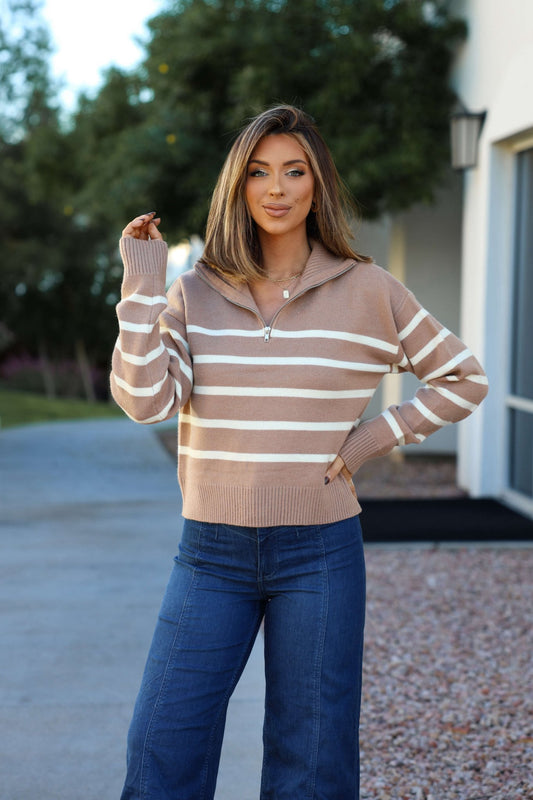 A woman wears a Taupe and Cream Striped Quarter Zip Pullover and blue jeans, smiling outdoors with one hand on her hip.