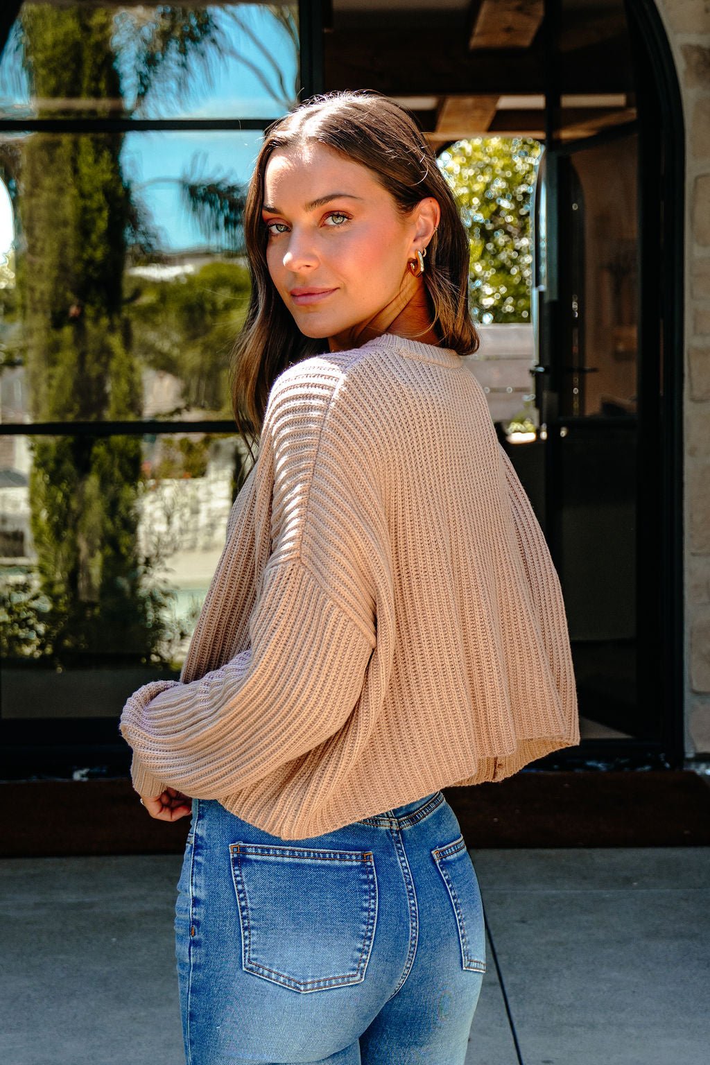 Woman in Taupe Boxy Ribbed Sweater - DOORBUSTER and blue jeans stands outdoors, looking over her shoulder at greenery and glass.