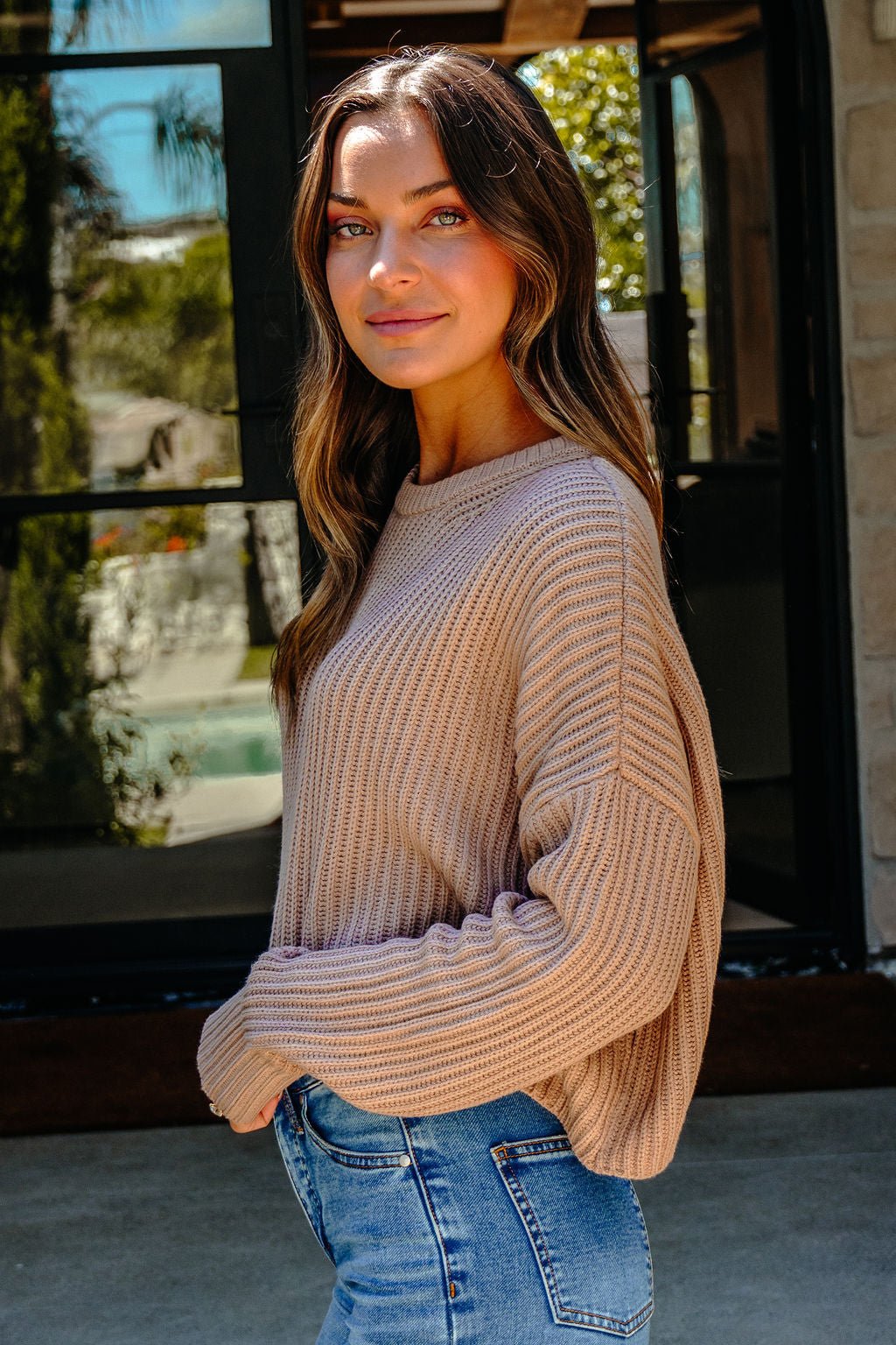 A woman in a Taupe Boxy Ribbed Sweater - DOORBUSTER and blue jeans stands outside, looking at the camera with a slight smile.