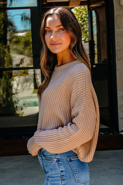A woman in a Taupe Boxy Ribbed Sweater - DOORBUSTER and blue jeans stands outside, looking at the camera with a slight smile.