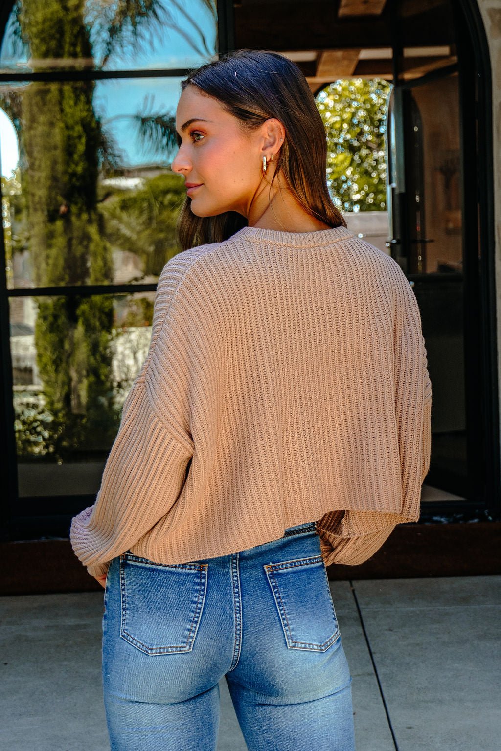 A woman in the Taupe Boxy Ribbed Sweater - DOORBUSTER stands outside near a window with greenery, facing away.