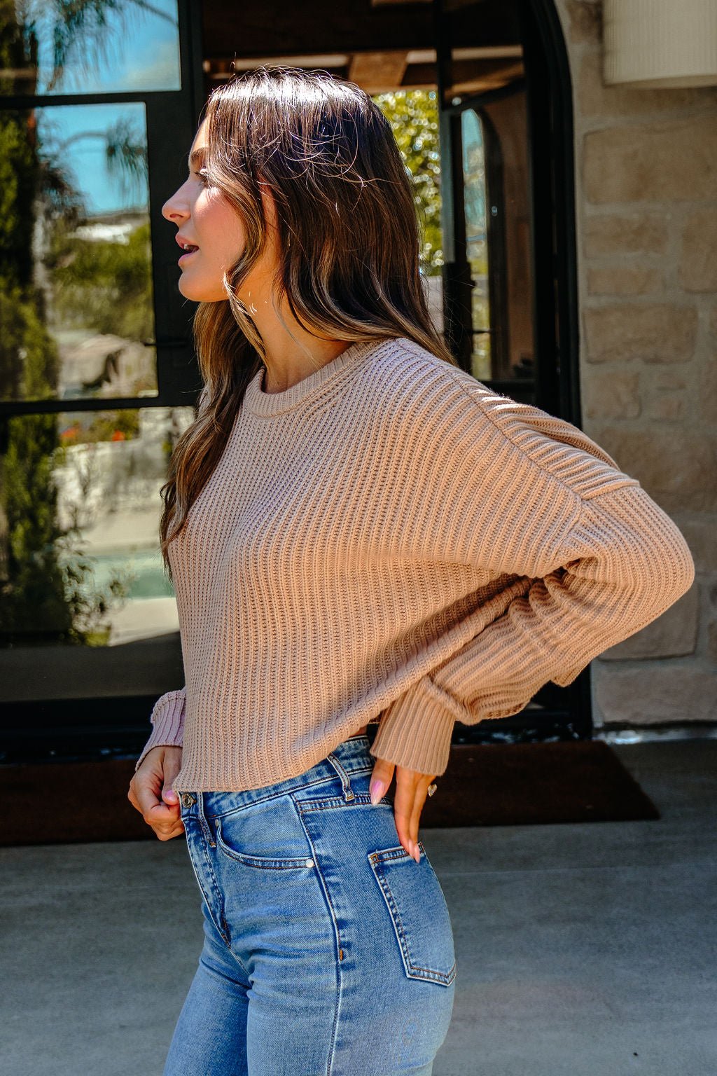 Woman in a Taupe Boxy Ribbed Sweater - DOORBUSTER and blue jeans stands outside, facing left with one hand in her pocket.