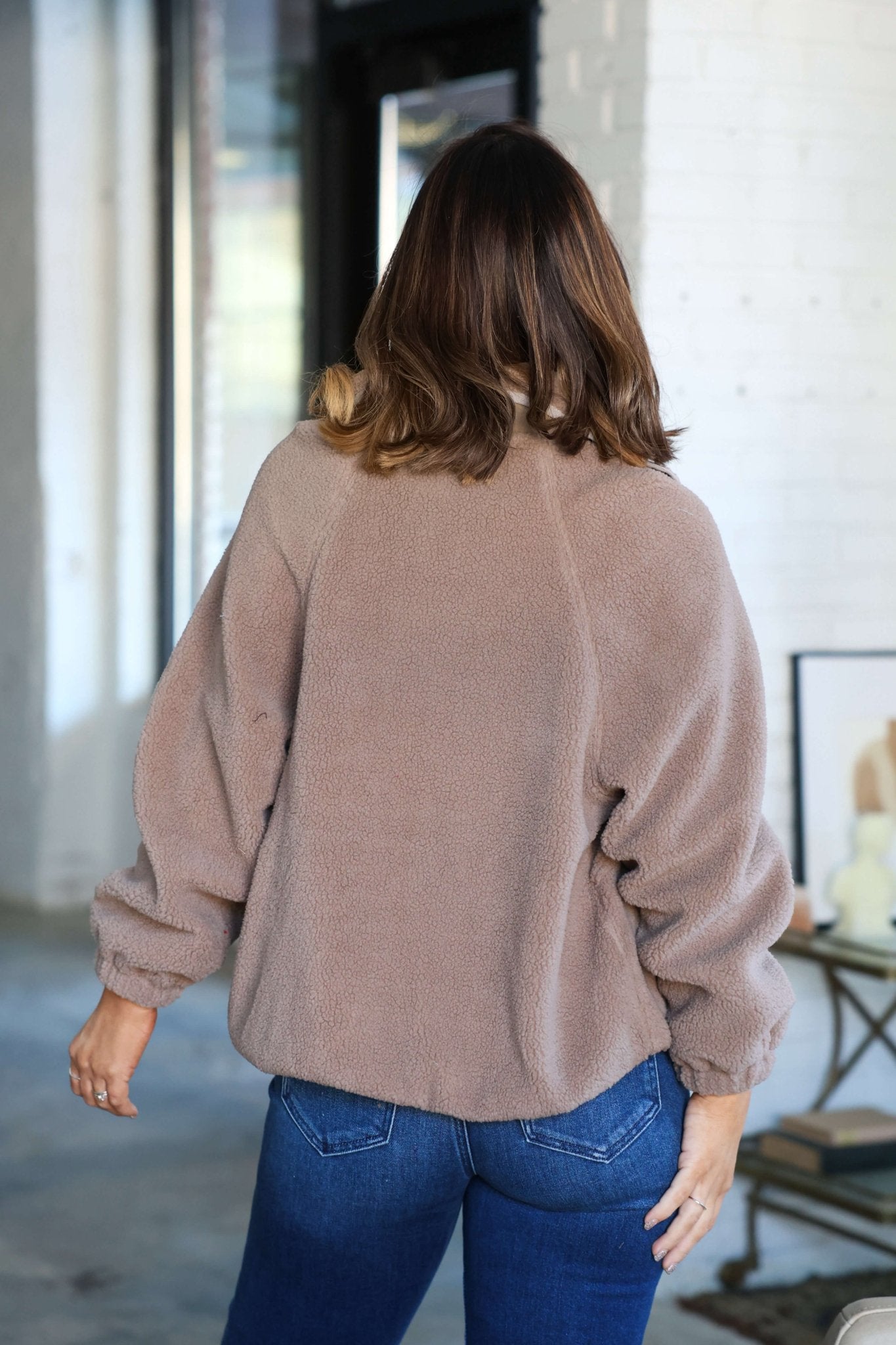 A woman with shoulder-length brown hair wears a Taupe Button Down Sherpa Jacket and blue jeans, facing away indoors.