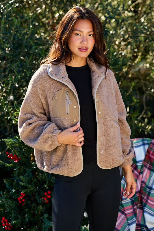 Woman in a Taupe Button Down Sherpa Jacket and black leggings stands outdoors by greenery and festive decorations, facing the camera.