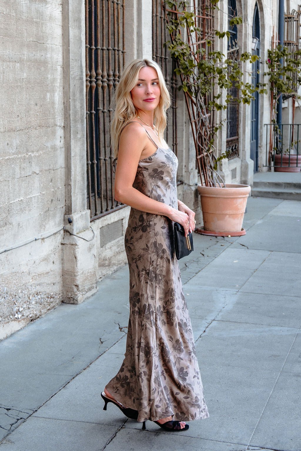 A woman in a Taupe Floral Print Satin Slip Dress and heels holds a black clutch by an iron-barred building window.