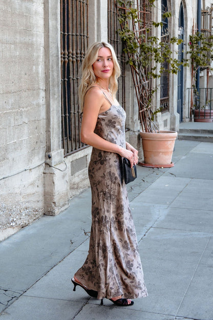 A woman in a Taupe Floral Print Satin Slip Dress and heels holds a black clutch by an iron-barred building window.