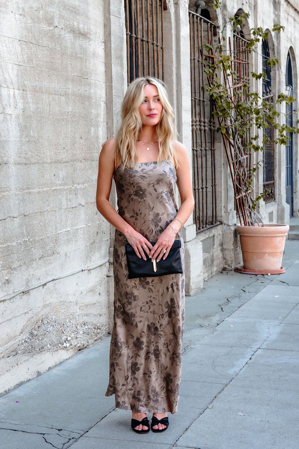 A woman in a Taupe Floral Print Satin Slip Dress holds a black clutch on a sidewalk beside a beige building with barred windows.