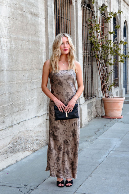 A woman in a Taupe Floral Print Satin Slip Dress holds a black clutch on a sidewalk beside a beige building with barred windows.