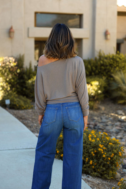 A woman stands outside on a path near bushes and yellow flowers, wearing a Taupe Off The Shoulder Crepe Top and blue jeans.