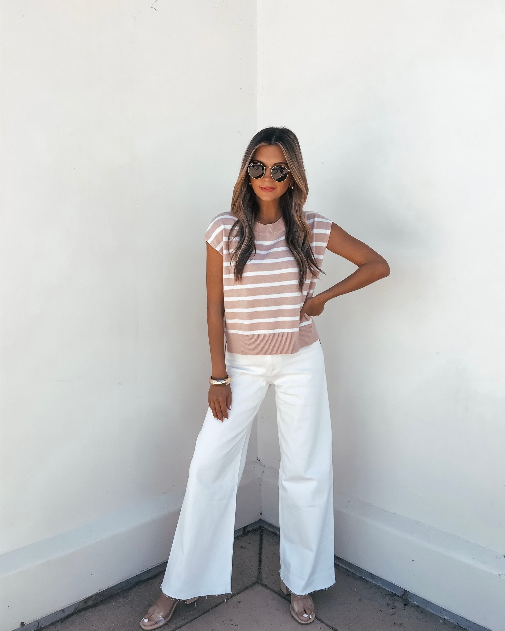 Woman in sunglasses wears a Taupe Striped Cap Sleeve Sweater, white wide-leg pants, and clear heels against a white wall.