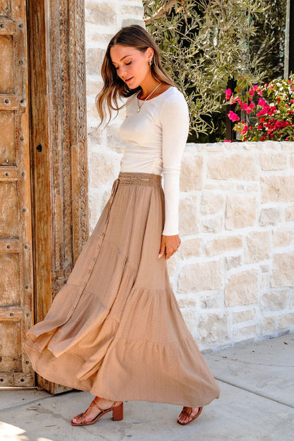 Woman in a white top and Taupe Striped Lace Trim Tiered Maxi Skirt stands outdoors by a stone wall and wooden door, flowers behind her.