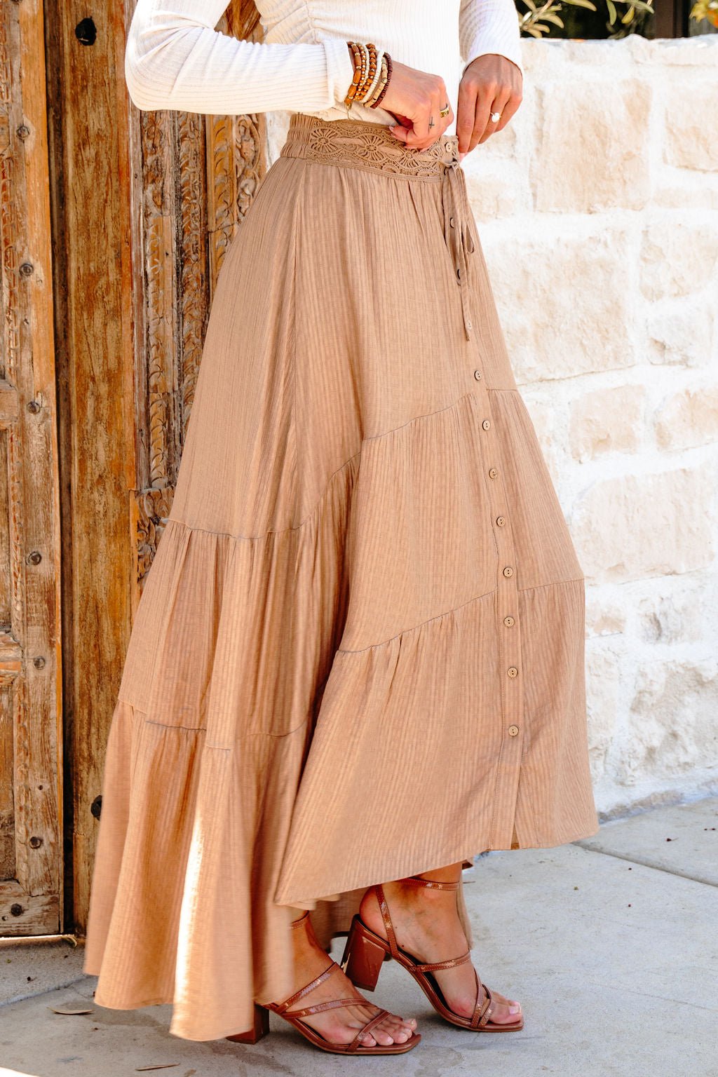 Woman in a Taupe Striped Lace Trim Tiered Maxi Skirt, sandals, and white top stands by a rustic wooden door and stone wall.