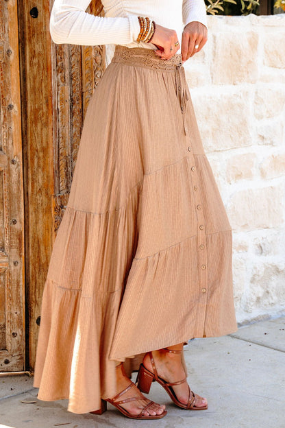 Woman in a Taupe Striped Lace Trim Tiered Maxi Skirt, sandals, and white top stands by a rustic wooden door and stone wall.