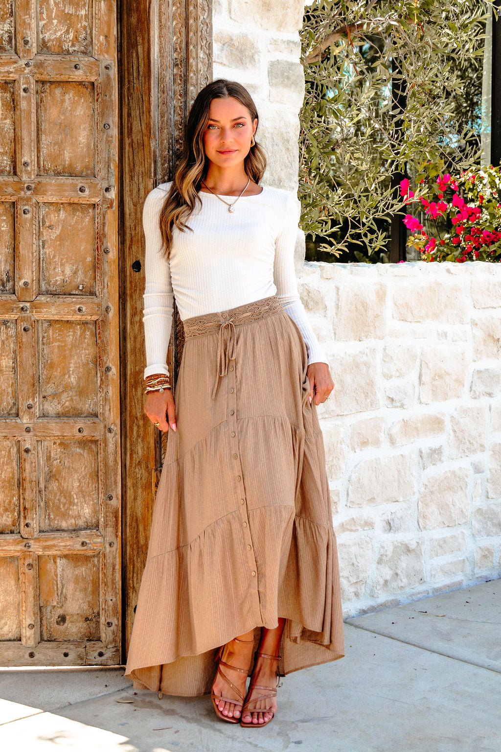 A woman smiles at the camera in a white long-sleeve top and a Taupe Striped Lace Trim Tiered Maxi Skirt by a rustic wooden door.