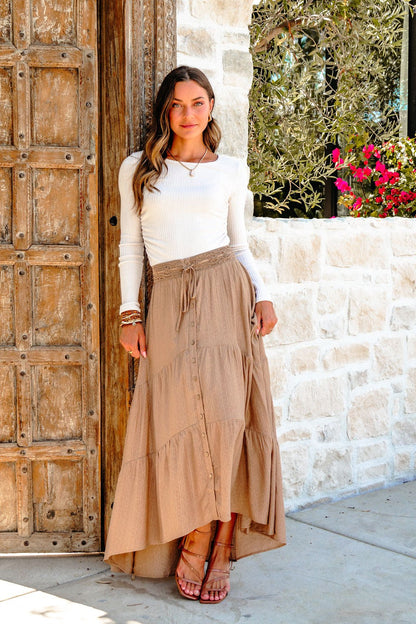 A woman smiles at the camera in a white long-sleeve top and a Taupe Striped Lace Trim Tiered Maxi Skirt by a rustic wooden door.