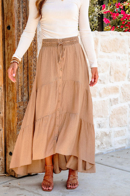 Woman wearing a Taupe Striped Lace Trim Tiered Maxi Skirt, white ribbed long-sleeve top, and tan sandals by a stone wall.
