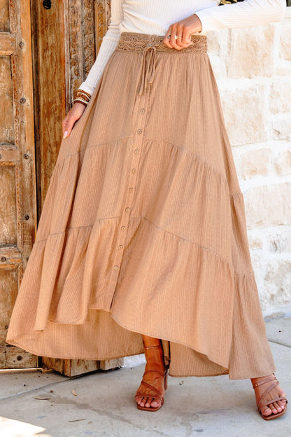 Wearing a Taupe Striped Lace Trim Tiered Maxi Skirt and brown sandals, a person stands by a wooden door and stone wall.