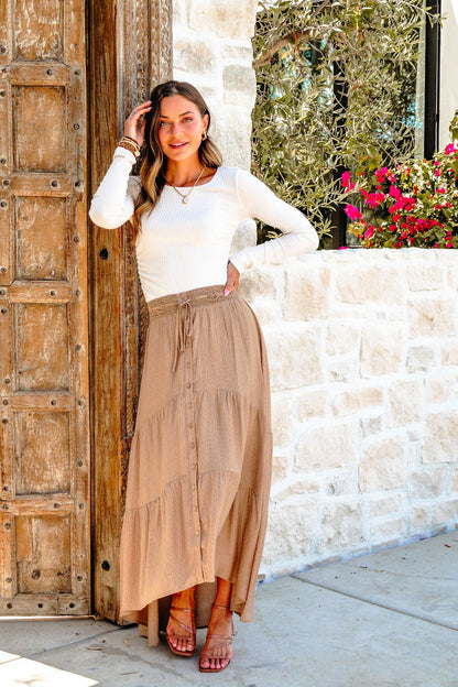 Woman in a white top and Taupe Striped Lace Trim Tiered Maxi Skirt smiles by a rustic door with stone walls and flowers.