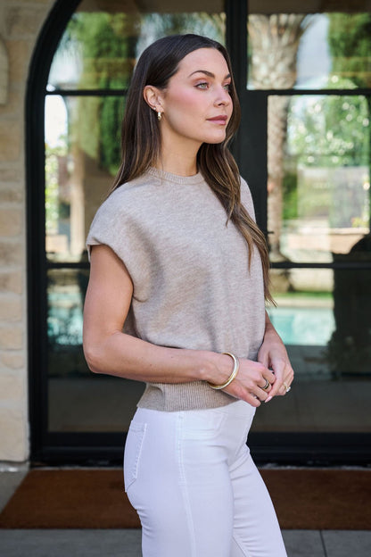 A woman with long brown hair wears a Taupe Two Tone Ribbed Sweater and white pants, standing near a glass door outdoors.