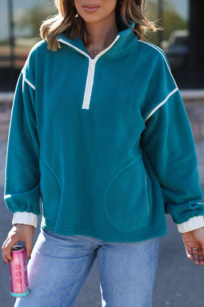 Woman in a Teal Contrast Half Zip Pullover and light blue jeans, holding a pink can outdoors.