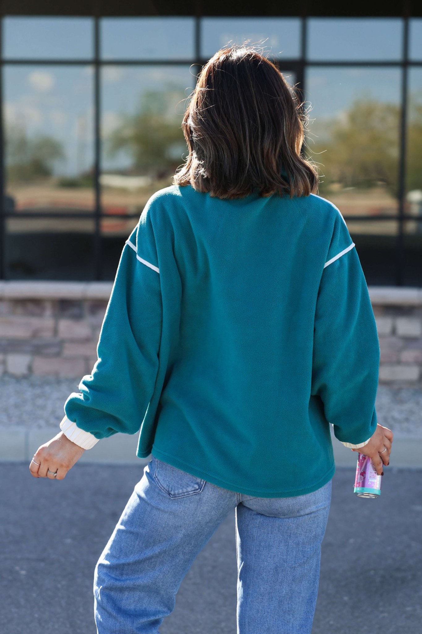 A person with shoulder-length hair stands outdoors facing away, wearing a Teal Contrast Half Zip Pullover and jeans, holding a can.