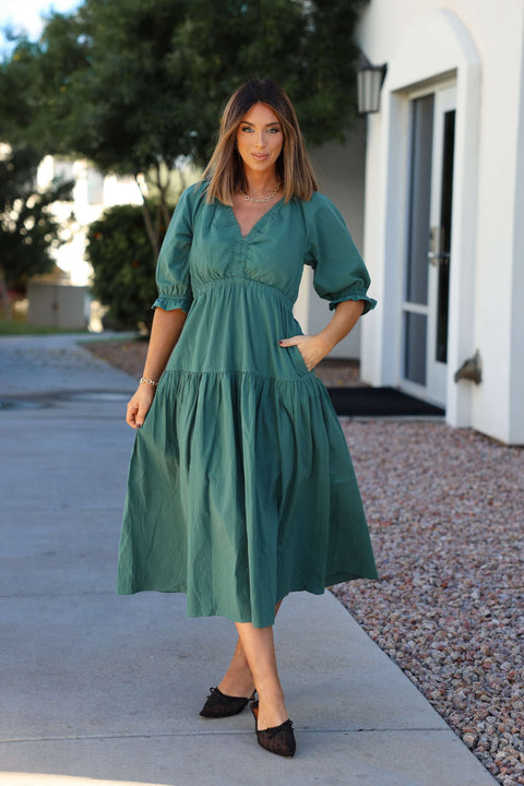 Woman in a Teal Puff Sleeve Tiered Midi Dress and black shoes stands outdoors near a white building and trees.
