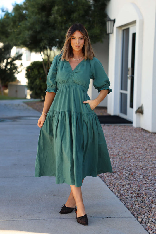 Woman in a Teal Puff Sleeve Tiered Midi Dress and black shoes stands outdoors near a white building and trees.