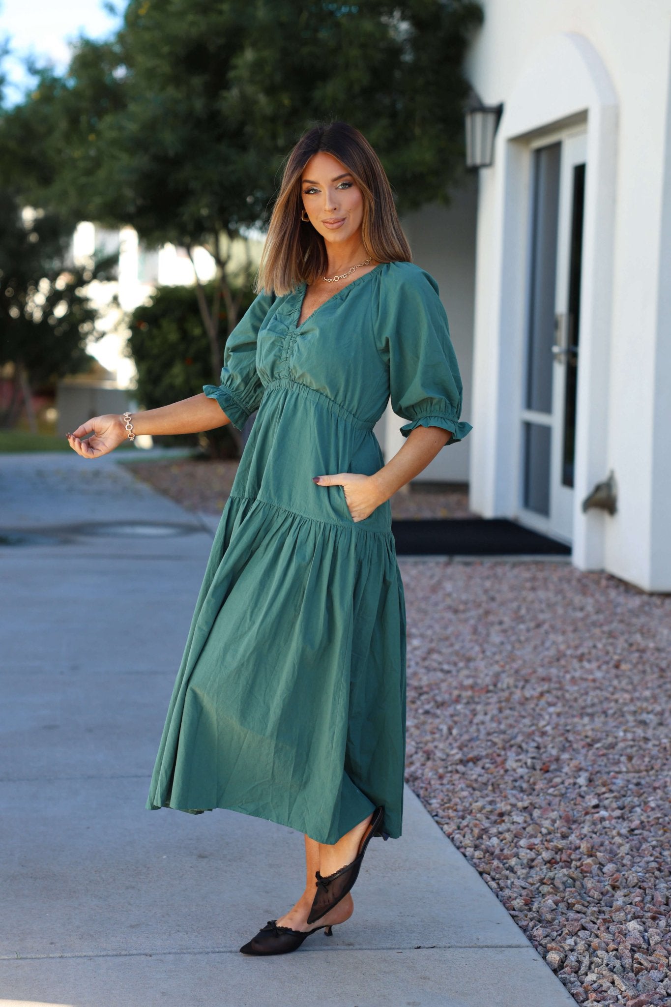 Woman wears a Teal Puff Sleeve Tiered Midi Dress while posing outdoors by a white building and landscaping.