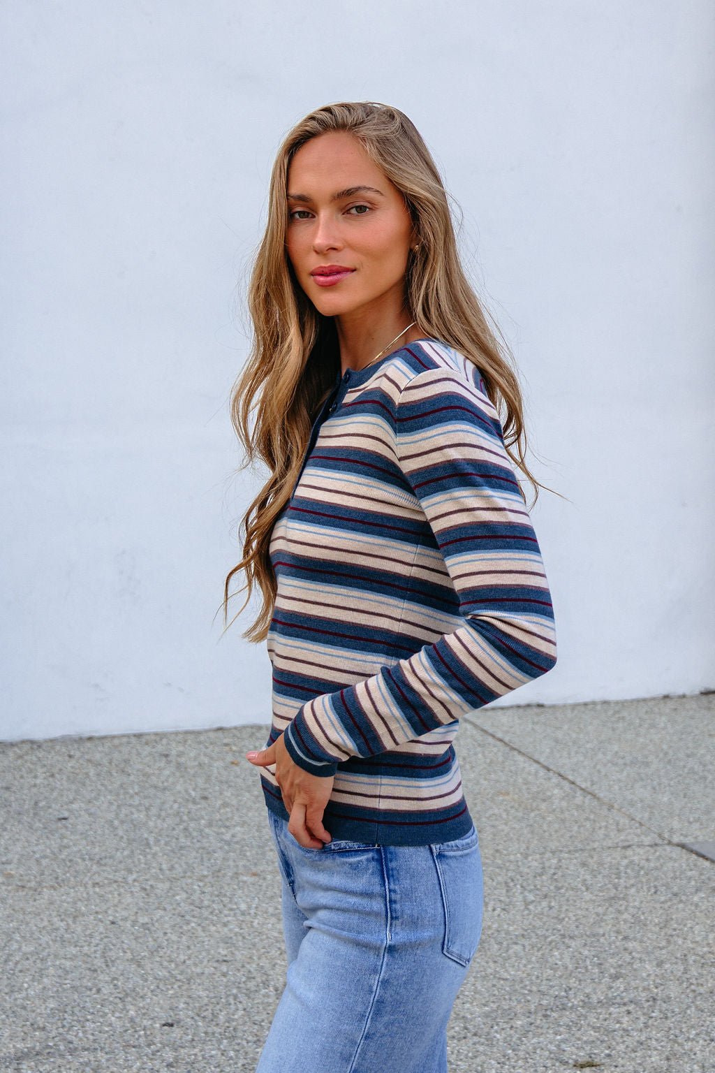 Woman with long hair models the Teal Striped Henley Long Sleeve Top and jeans outdoors against a plain white wall, facing the camera.