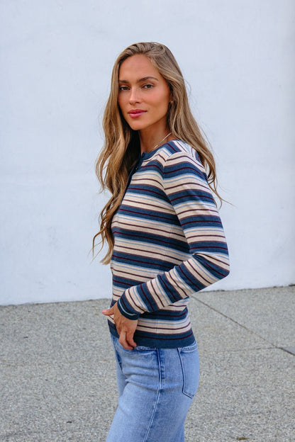 Woman with long hair models the Teal Striped Henley Long Sleeve Top and jeans outdoors against a plain white wall, facing the camera.