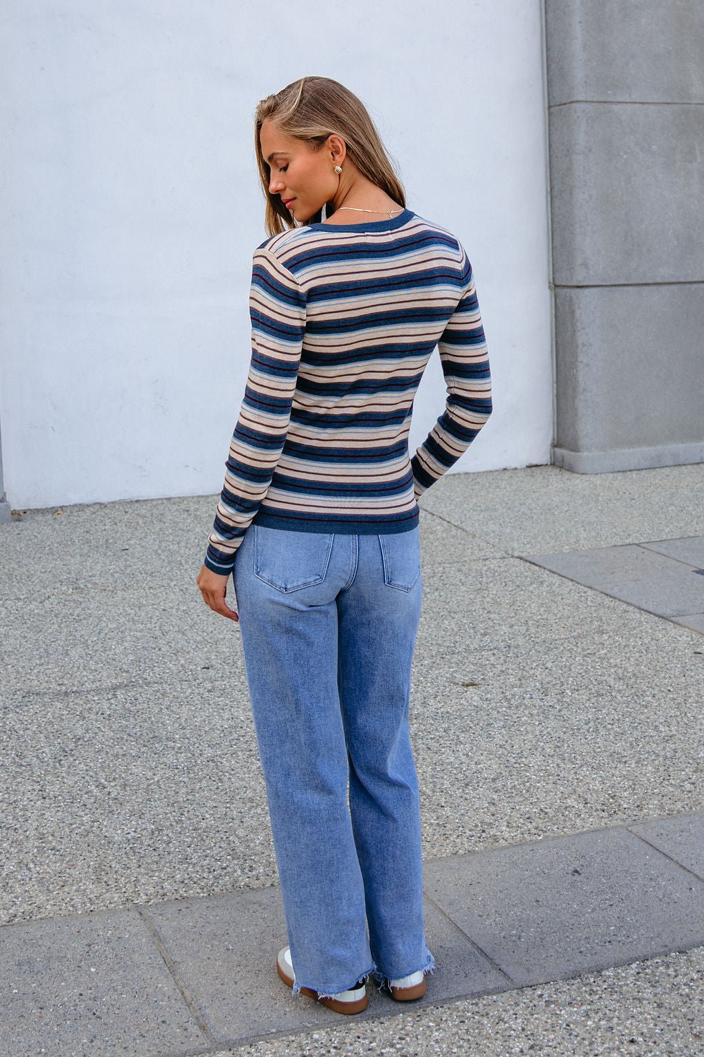 Woman outdoors in a Teal Striped Henley Long Sleeve Top and light blue jeans, standing on stone pavement, facing away.