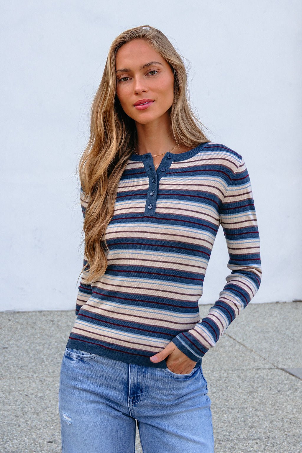 Woman with long blonde hair models the Teal Striped Henley Long Sleeve Top and jeans, standing against a plain background, hand in pocket.